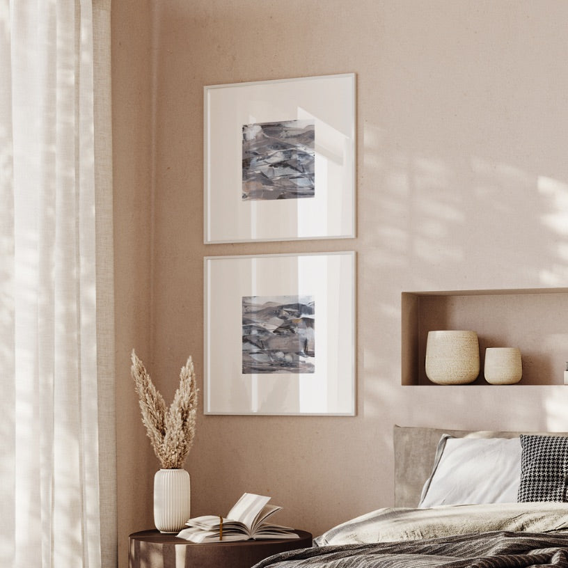 Two framed abstract art prints with gray, blue, and white tones hang on a warm beige wall above a bed in a softly lit bedroom. A small side table with a vase of dried grass and an open book sits below.